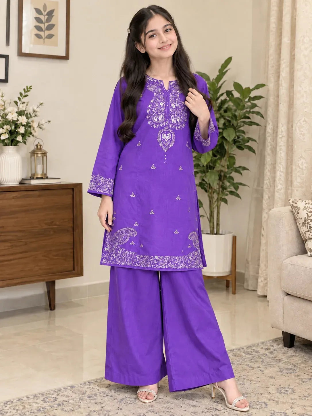 A young girl modeling a purple three-piece salwar kameez suit with white embroidery, standing in a decorated living room.