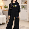 A young girl stands in a cozy living room wearing a matching black two-piece pajama set with colorful cartoon butterflies and the words 'hello winter' on the sweatshirt and pants.