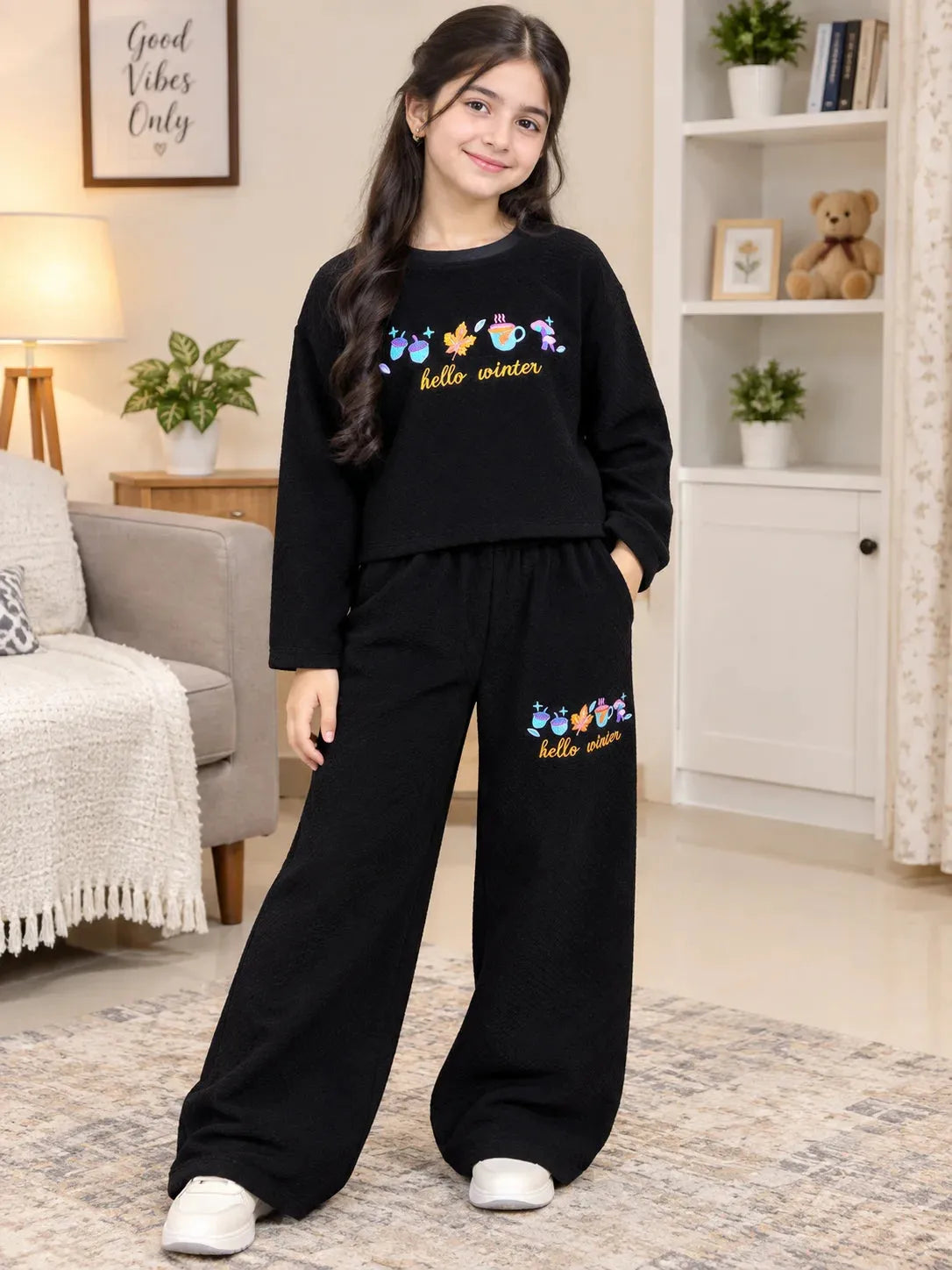 A young girl stands in a cozy living room wearing a matching black two-piece pajama set with colorful cartoon butterflies and the words 'hello winter' on the sweatshirt and pants.