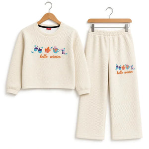 A two-piece children's lounge set consisting of a cropped long-sleeve top and matching pants, both in a textured cream waffle fabric. The set features a colorful graphic of winter-themed icons (snowman, scarf, hat, gloves, sunglasses) above the text 'hello winter' in orange.