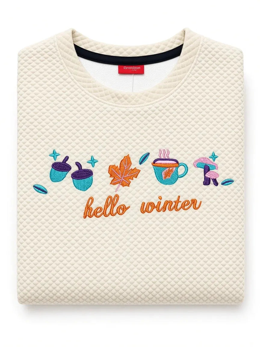 A folded cream-colored quilted sweatshirt featuring colorful embroidered illustrations of acorns, a maple leaf, a coffee cup, a mushroom, and the text 'hello winter' in orange.