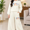 A young girl stands in a living room wearing a cream-colored two-piece pajama set with a textured waffle pattern. The set includes a long-sleeve sweatshirt and matching wide-leg pants, both featuring a colorful fish graphic and the words 'hello winter' on the front.