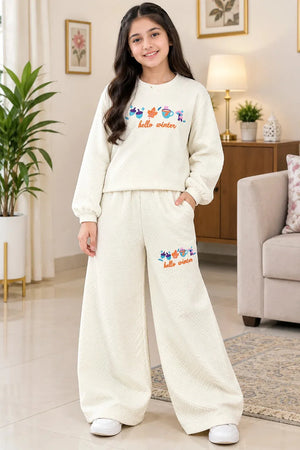 A young girl stands in a living room wearing a cream-colored two-piece pajama set with a textured waffle pattern. The set includes a long-sleeve sweatshirt and matching wide-leg pants, both featuring a colorful fish graphic and the words 'hello winter' on the front.