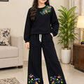 A young girl stands in a living room wearing a black two-piece loungewear set with colorful floral embroidery on the chest and lower legs. The set includes a long-sleeve top and matching wide-leg pants with a drawstring waist.