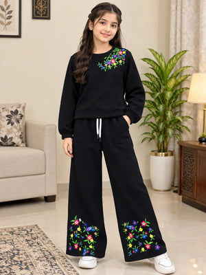A young girl stands in a living room wearing a black two-piece loungewear set with colorful floral embroidery on the chest and lower legs. The set includes a long-sleeve top and matching wide-leg pants with a drawstring waist.