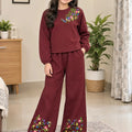 A young girl stands in a living room wearing a matching two-piece lounge set. The set consists of a textured, long-sleeve crewneck top and wide-leg pants in a deep burgundy color, both decorated with colorful floral embroidery.