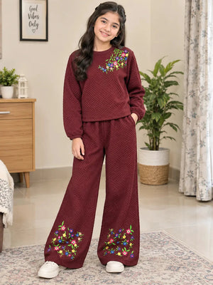 A young girl stands in a living room wearing a matching two-piece lounge set. The set consists of a textured, long-sleeve crewneck top and wide-leg pants in a deep burgundy color, both decorated with colorful floral embroidery.