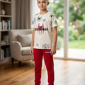 A young boy wearing a Spider-Man themed two-piece outfit, featuring a cream-colored t-shirt with a red and black spider logo and blue spider prints, paired with bright red sweatpants and white sneakers, standing in a modern living room.